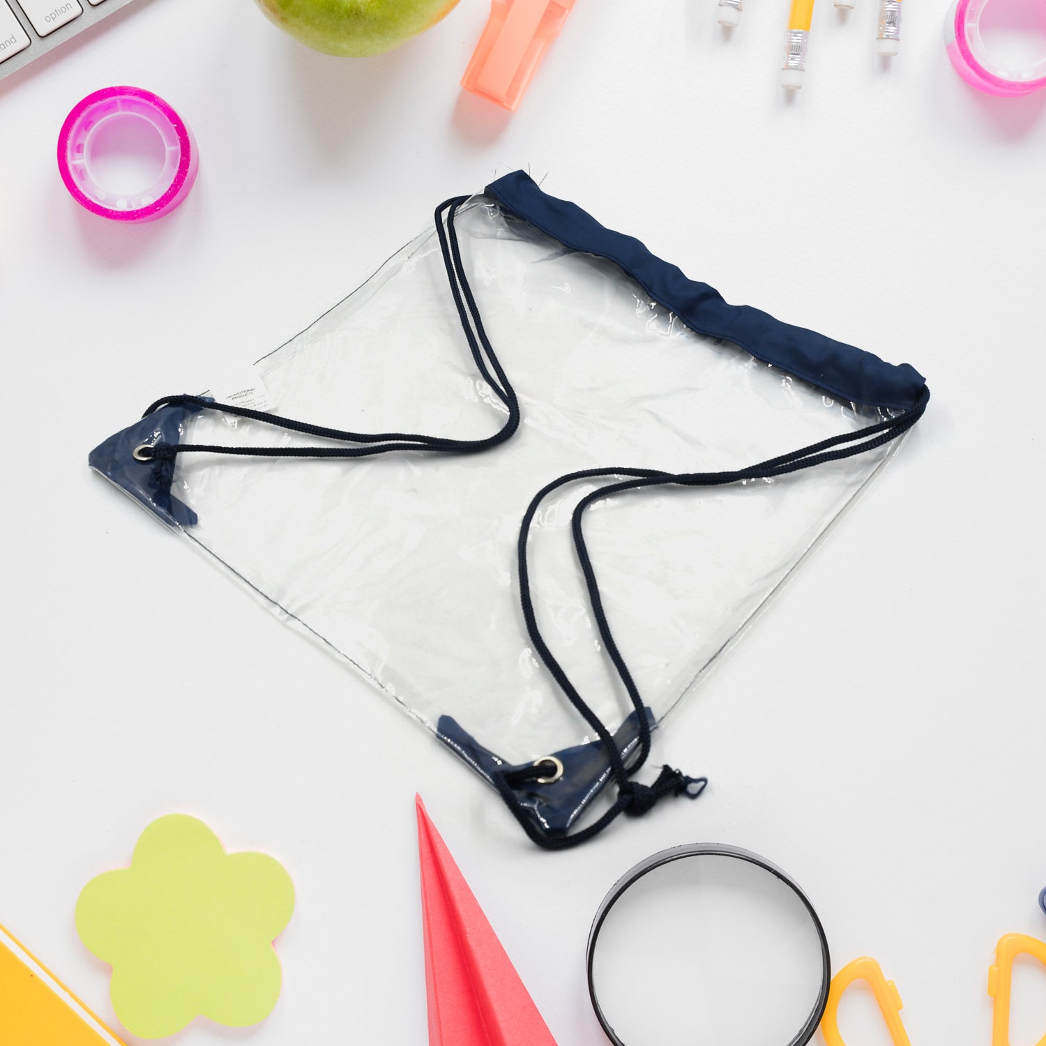A clear drawstring bag with navy blue accents is centered on a white desk with various office supplies scattered around it.