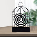 A black iron birdcage-style holder with a burning mosquito coil inside sits on a light wooden table.