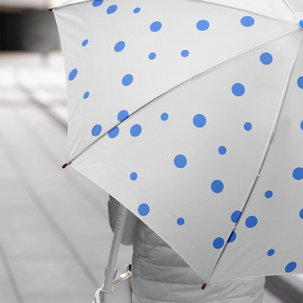 A person holds an open white umbrella with blue polka dots, with a gray background and a hint of a jacket.