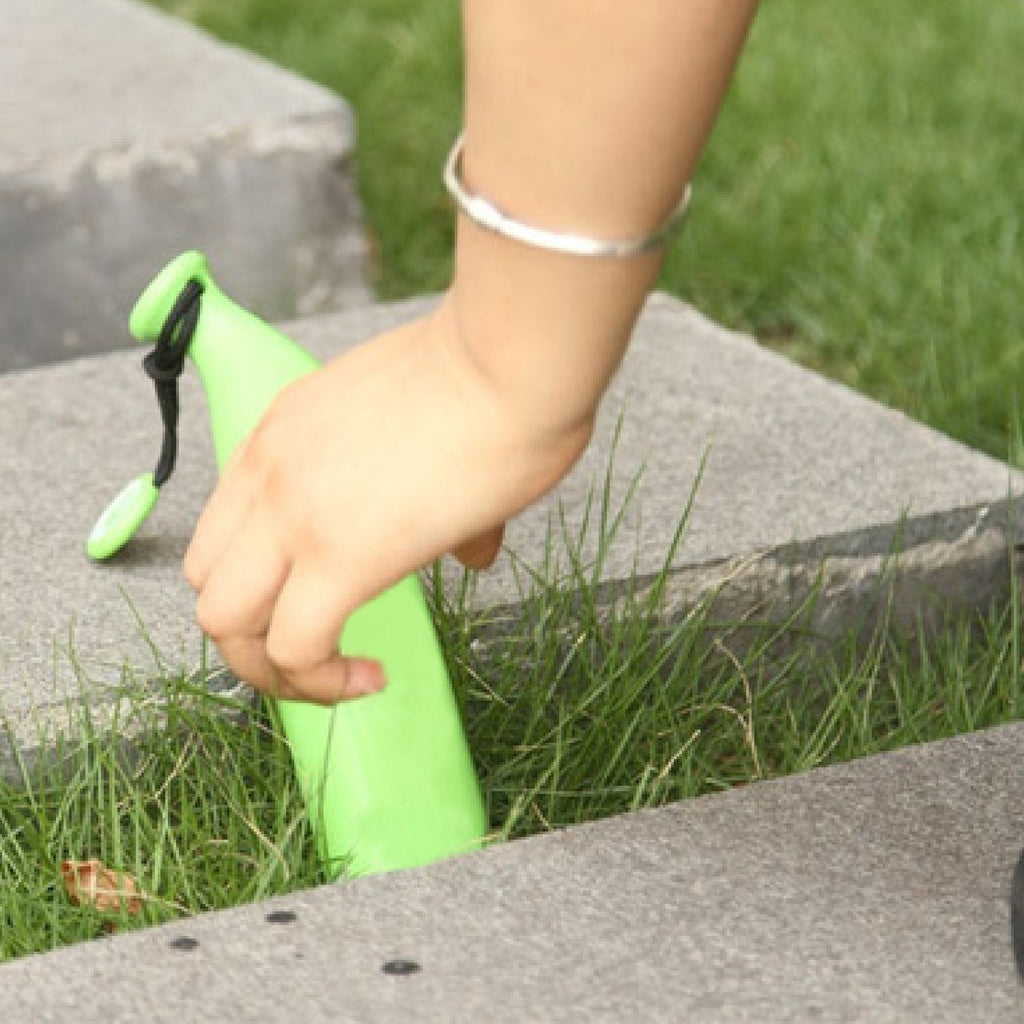 A hand reaches for a green banana-shaped umbrella case resting in green grass between concrete slabs.