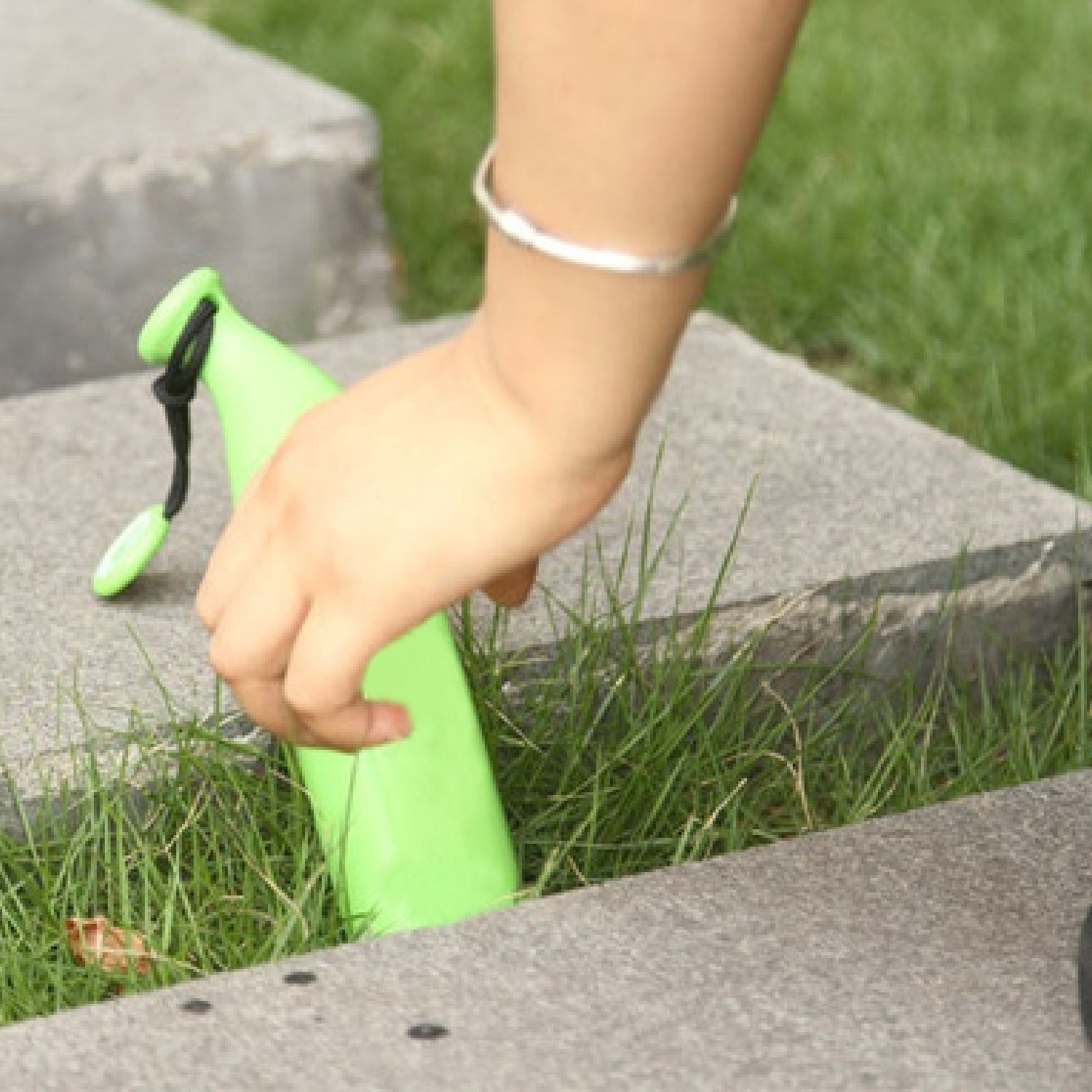 A hand reaches for a green banana-shaped umbrella case resting in green grass between concrete slabs.
