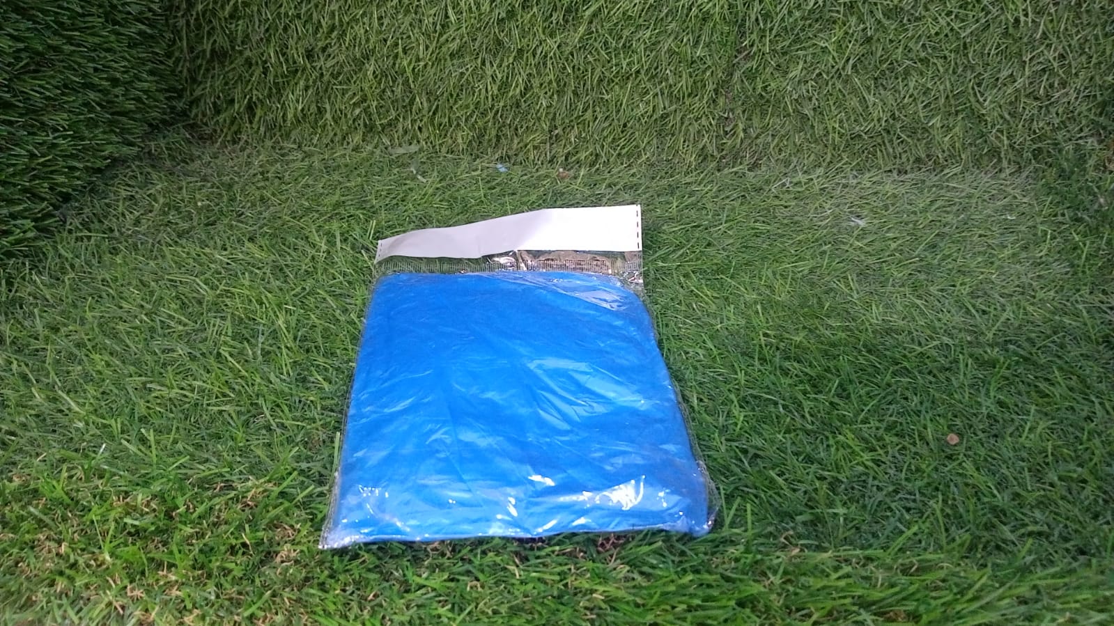 A small, clear plastic package containing a folded blue poncho rests on a vibrant green grass lawn.